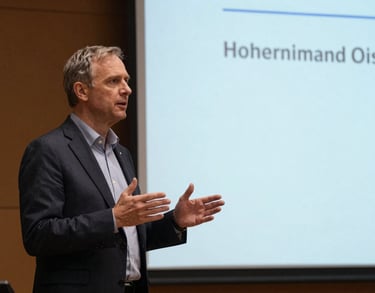 A speaker on stage at an educational conference, gesturing towards a presentation screen. North American / International academic setting.
