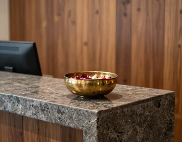 A serene office reception area with a modern stone desk, a South Asian decorative brass bowl with floating flower petals, and warm wooden wall panels.