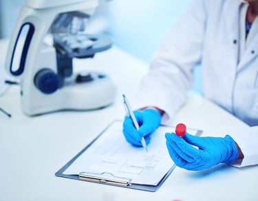 Scientist in white coat and blue gloves holding a sample vial near a microscope and clipboard.