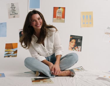 Smiling woman in a white linen shirt and jeans sitting in front of a wall with skincare mood board inspiration.