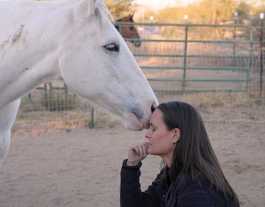 A white horse gently touches its nose to a woman's forehead during an equine therapy session.