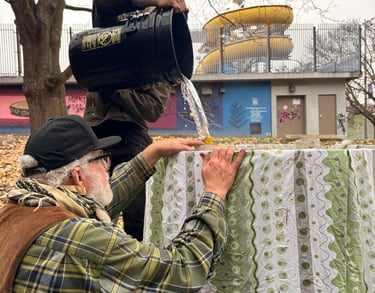 A man pours water from a black bucket over a fabric-covered structure in an urban park.