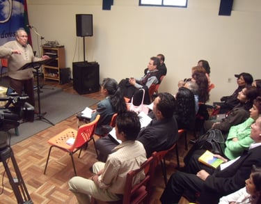 An elderly man speaking at a lectern during a community meeting or seminar with an attentive seated audience.