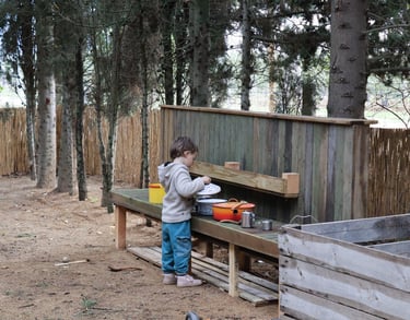 Infant jugant a la cuineta exterior de fusta a l’escoleta, amb estris i olles