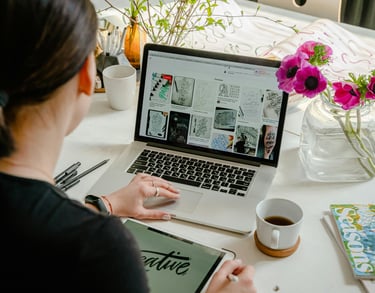 A female graphic designer using a laptop and tablet at a white desk with flowers and coffee.