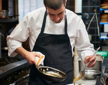 Chef wearing an uniform, pouring sauce from a pan into a bowl of clams in a professional kitchen.