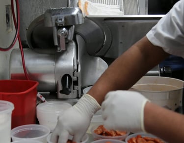 Gloved hands placing sliced chorizo into containers in a professional kitchen.