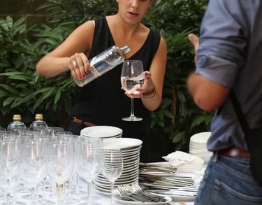 Woman dressed in black pouring water into a glass next to a table set with plates and glasses in a restaurant.
