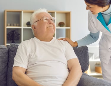 un homme âgée assis sur un fauteuil la main d'une soignante sur l'épaule gauche.