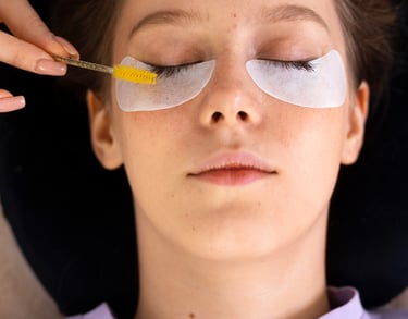 A beauty technician uses a yellow spoolie brush to groom eyelashes during a professional eyelash extension treatment.