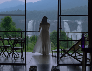 Woman in white dress standing on balcony overlooking Athirappilly Falls, framed by glass doors.