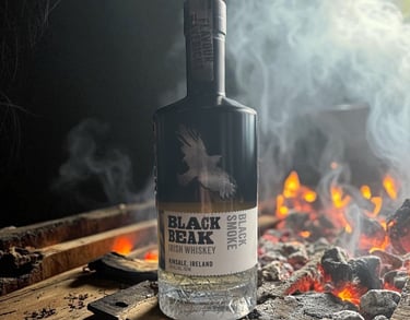 A bottle of Black Beak Black Smoke Irish Whiskey sits on wood planks next to a smoky charcoal fire.