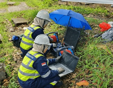 Two engineers in safety gear perform a geophysical survey using a laptop and seismic equipment outdoors.