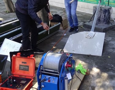Technicians operate a borehole inspection camera with a motorized winch and control box for geological survey.