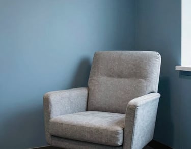 A comfortable, modern armchair in a light-filled corner of an office with soft steel blue walls, representing a safe space for individual therapy.