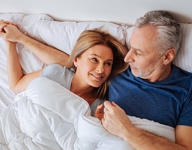 A smiling mature woman and man lying in bed holding hands and looking at each other affectionately.