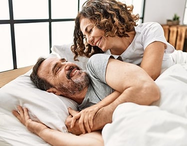 A joyful middle-aged man and woman with curly hair laughing and embracing while lying in bed