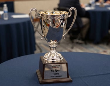 A close-up of a silver championship trophy sitting on a table with a dark navy blue tablecloth. Soft light reflects off the metal. North American / US Southern collegiate athletic banquet setting.