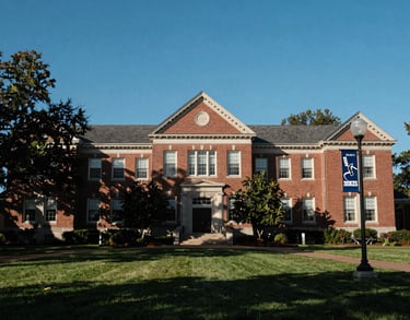 Photography of a large university campus building with traditional Southern architecture, surrounded by green lawns under a clear blue sky. A banner for the hockey program is visible on a nearby lamp post.