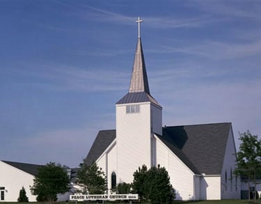 Peace Lutheran Church building featuring a white exterior with a tall steeple against a clear blue sky.