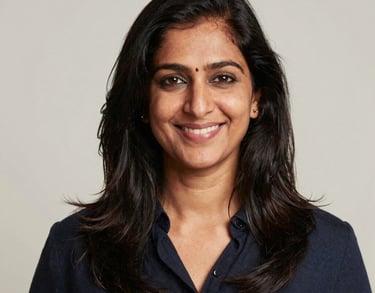 A professional studio portrait of an approachable South Asian woman smiling, set against a neutral off-white background. Professional lighting highlighting a trustworthy and warm persona.