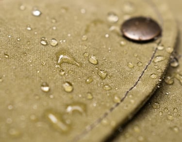 Water droplets beading on a waterproof olive green waxed canvas jacket with a metal button.