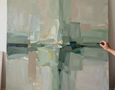 Photography of a large abstract painting in progress, with soft washes of sage green and taupe. The artist's hand is visible in the corner, holding a charcoal stick, in a sun-drenched North American studio.