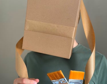 Photo shows a man with cardboard boas on his head covering his face. Man holds paint brushes.