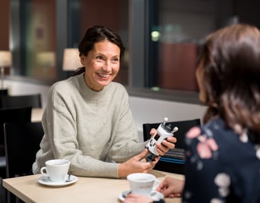 Woman in cafe, holding an iCare HOME2 and discussing with her friend, about measuring her own eye pressure IOP at home.