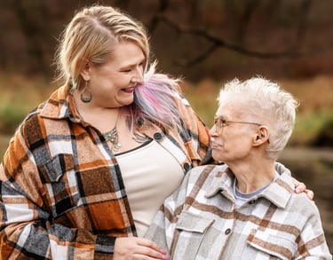 A blonde woman with pink hair highlights and an older woman wearing flannel shirts smiling outdoors.