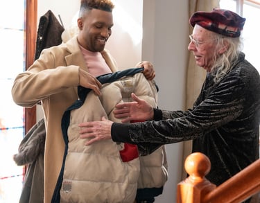 A young man in a tan overcoat helps a senior man put on a winter puffer jacket at home.