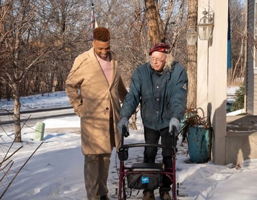 A young caregiver assists a senior man using a walker on a snowy sidewalk outside a home.