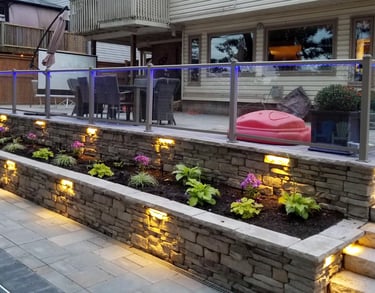 Illuminated stacked stone retaining wall with garden bed and glass railing on a backyard patio.