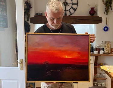 The artist holds a vibrant red sunset oil painting on canvas in a gold frame.