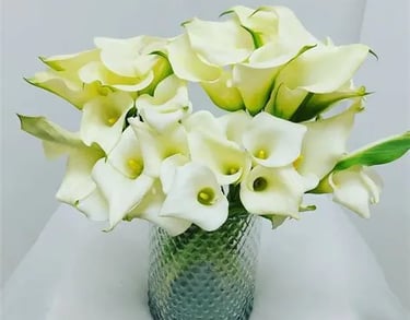 A large bouquet of white calla lilies with yellow centers in a textured grey-green glass vase.