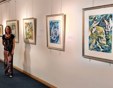 Artist standing next to a wall of colorful abstract framed paintings in an art gallery exhibition.