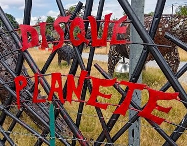 Red metal letters spelling Disque Planete on a black lattice fence near metal scrap sculptures.