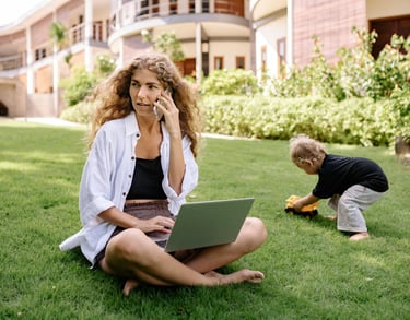 Woman working remotely on laptop and phone in a park while a child plays, symbolizing flexib