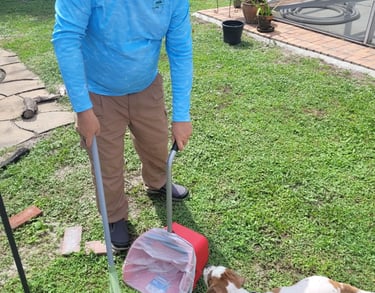 Robert using a sanitized scooper to remove waste from a customers back yard, small dog watching him