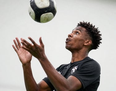 Action shot of a young male player of African descent, aged 15, performing a header. Sharp focus, clean white background of a professional facility wall. Intense commitment in his eyes.