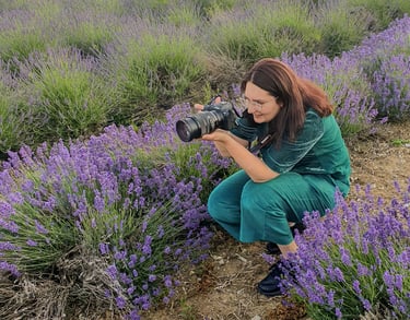 A female photographer holding a camera crouches in a blooming purple lavender field.