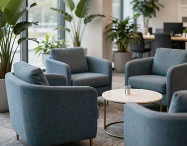 A bright collaboration zone with comfortable lounge chairs in slate blue, surrounded by indoor plants and natural light in a US corporate setting.
