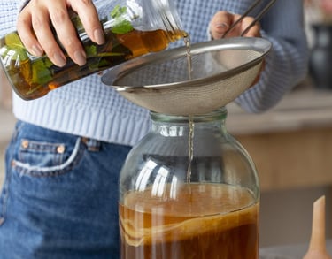 Una mujer vertiendo té casero en un frasco de fermentación de kombucha de vidrio con un SCOBY.