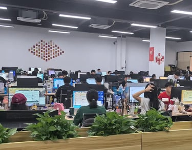 Employees working at computer desks in a modern open-plan office with green plants.