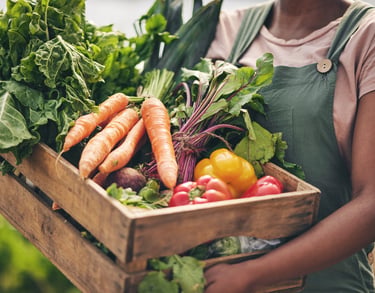 A farmer holding a wooden crate filled with fresh organic vegetables like carrots, peppers, and leafy greens.