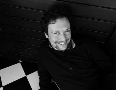 A smiling man with a beard wearing a black shirt in a black and white portrait.