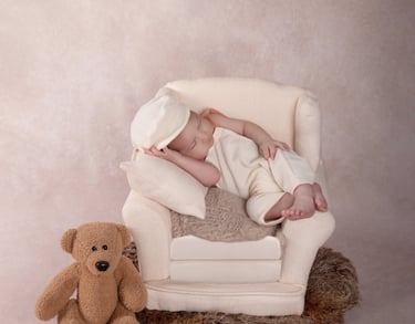 un bebé dormido en un pequeño sillón, al lado se encuentra un osito de peluche