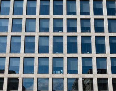 Photography of an official building facade with modern glass architecture reflecting a blue sky. Clean, authoritative, and professional. Medium blue and off-white tones. Global / Spanish-speaking.