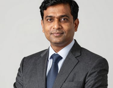 A professional studio portrait of a South Asian male educator in formal attire. Sharp lighting, clean professional aesthetic against a light background.