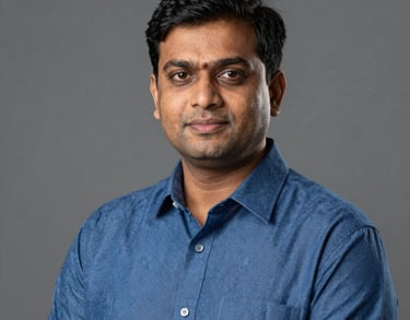 A professional studio portrait of a South Asian male teacher in a blue formal shirt. He looks approachable and authoritative. Neutral gray background.